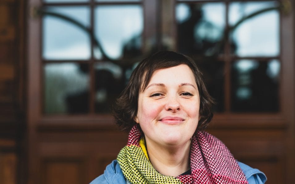 Josie Long smiling in front of wooden doors, wearing a colourful scarf.