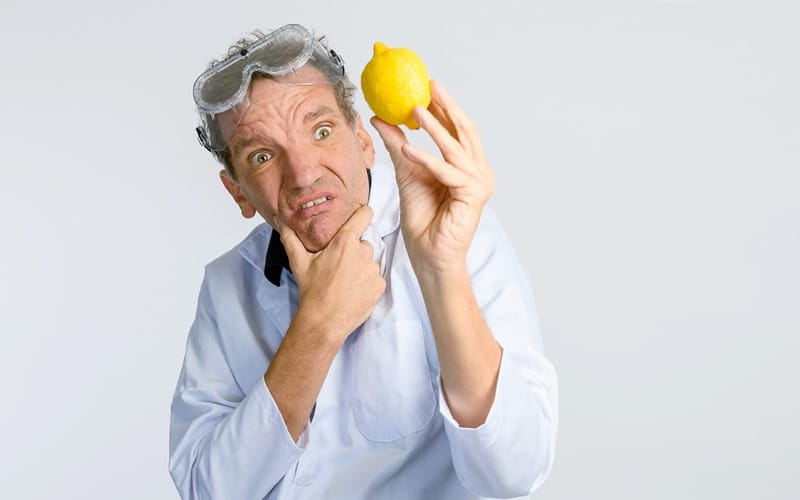 A man in a lab coat and goggles peers suspiciously at a lemon.