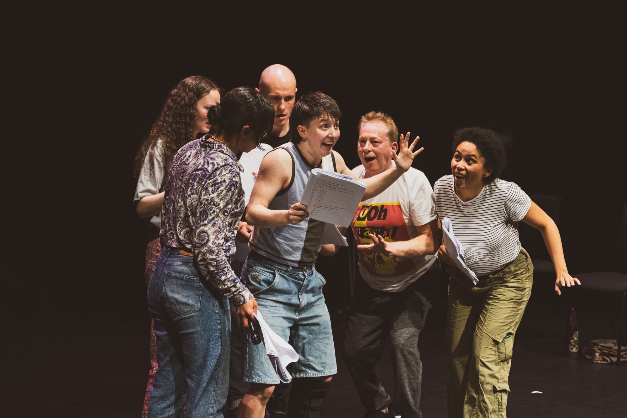 A group of performers on stage, huddled around a script and laughing