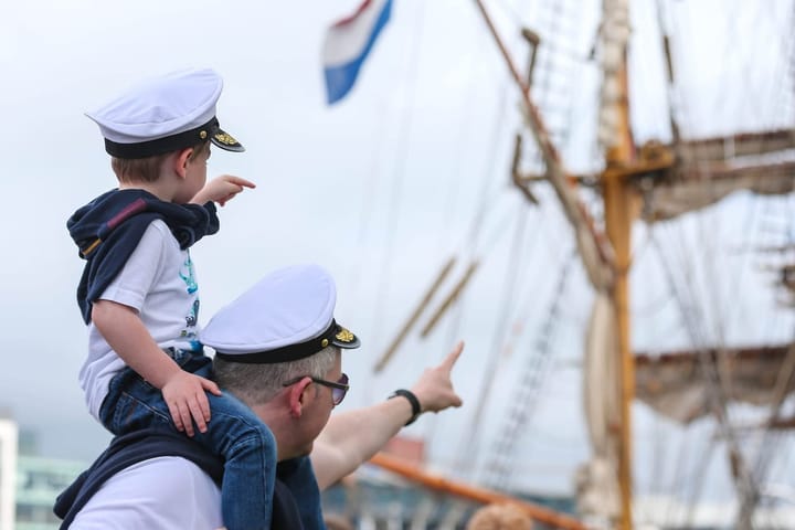 A man with a child on his shoulders, both wearing sailor hats, point excitedly towards a tall ship docked nearby, with rigging and a flag visible in the background.