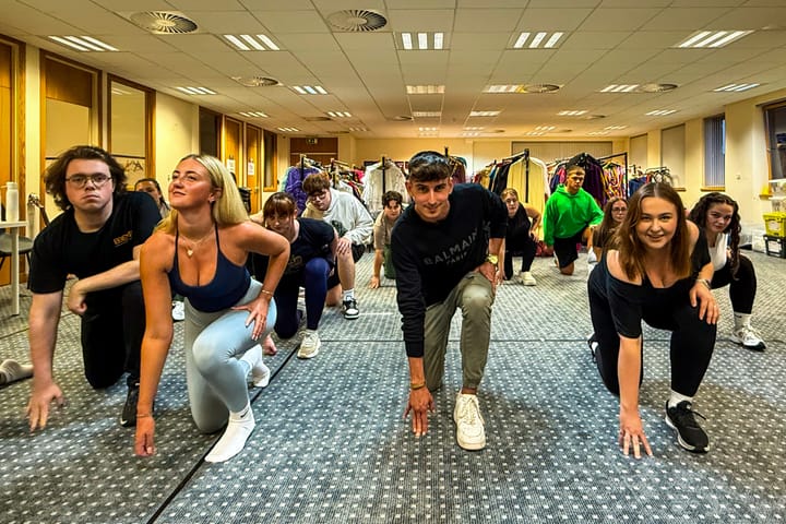 Performers crouch in formation mid-rehearsal in a costume-filled practice space.