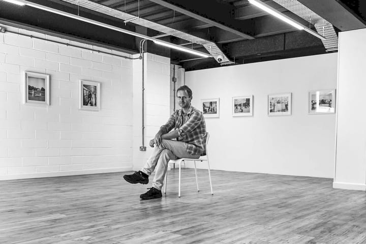 B&W portrait of Lee Garson seated in a gallery with framed photos on the wall.
