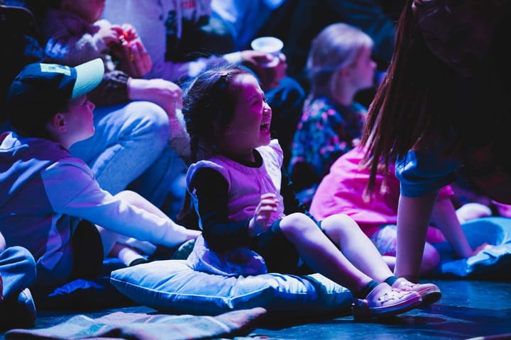 Young girl laughing on a cushion at a children’s show, surrounded by other kids.
