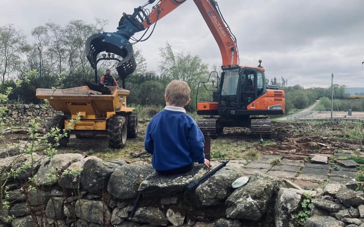 A child in a blue school jumper sits on a stone wall watching an orange digger.