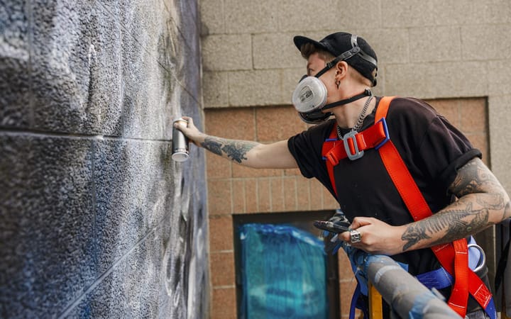 An artist in a respirator mask and safety harness spray paints a large mural on granite.