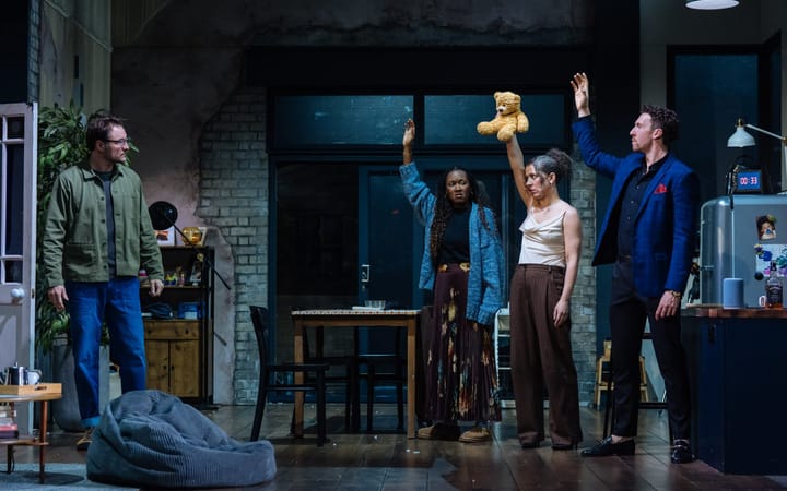 Four actors on stage, two with arms raised, holding a teddy bear.