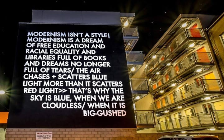 Illuminated text artwork by Robert Montgomery on a building wall at night