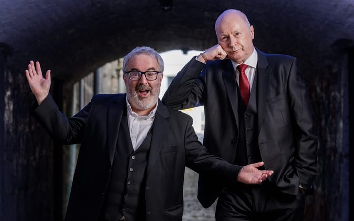 Two men in dark suits pose theatrically in a stone archway