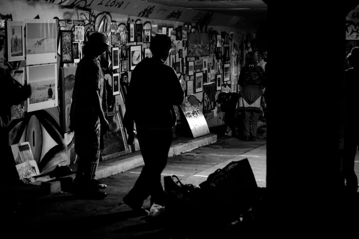 Silhouetted visitors view framed artworks on a graffiti-covered underground car park wall