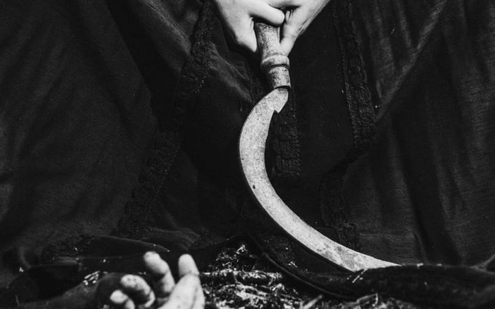 A rusted sickle curves across dark fabric, one hand on the handle, another partially buried in the ground below