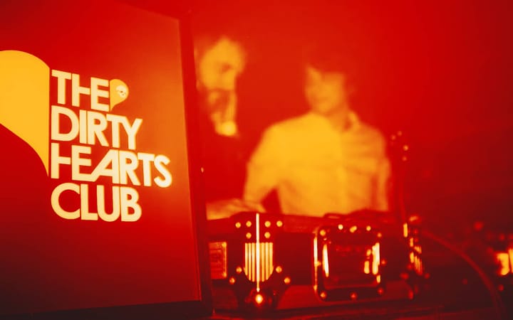 The Dirty Hearts Club sign lit up red beside a DJ silhouetted at the decks