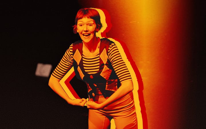 Comedian Elf Lyons performing on stage in a striped top and diamond waistcoat