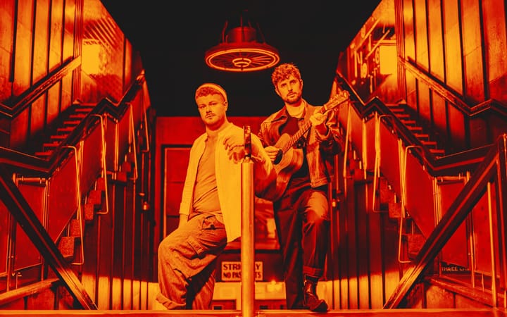 Jamie Rea and Dylan Wood pose on a staircase in a red-toned publicity shot for The Singer.