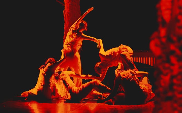 Dancers in pale costumes move together under deep red stage lighting.